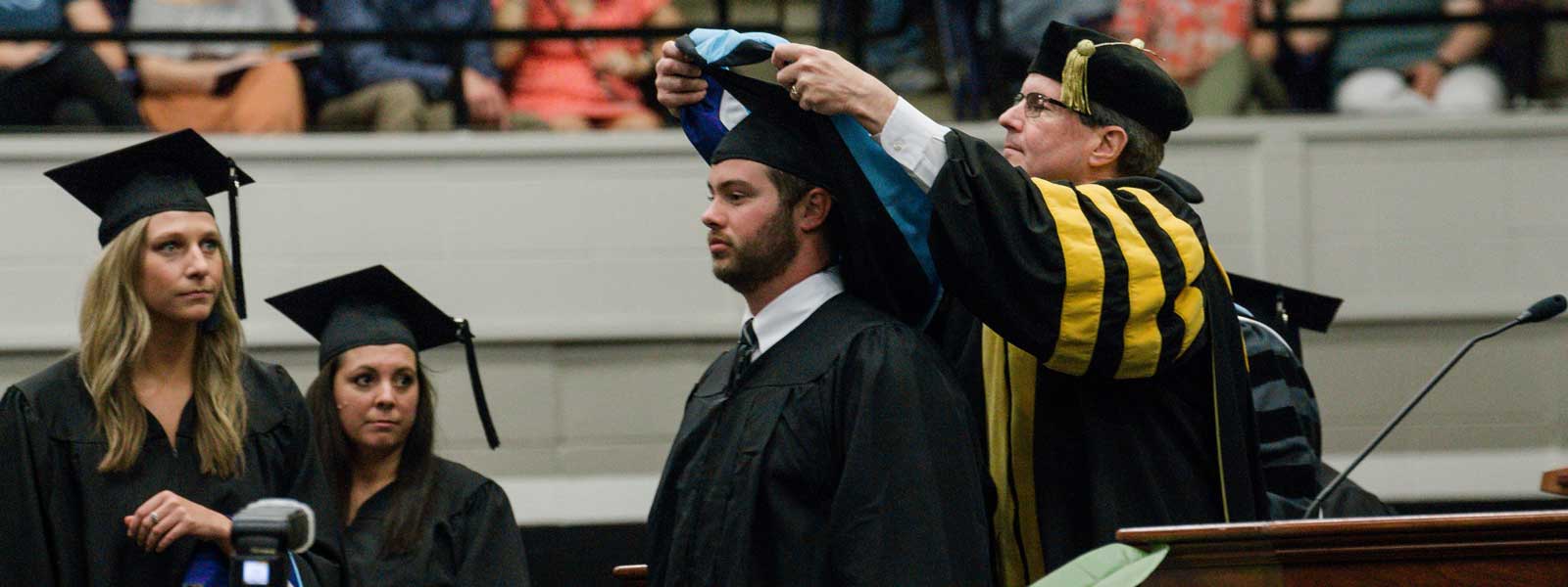 college dean put hoods around neck of graduate while other graduates watch and wait their turns