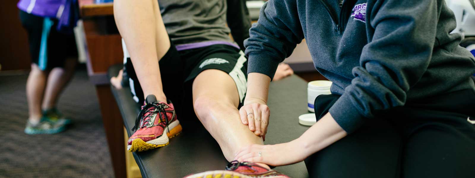 student athletic training performs muscle therapy exercise on athlete's leg
